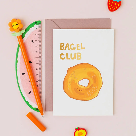Bagel Club greeting card featuring a hand drawn smiling sesame bagel with gold foil lettering on a contemporary white background.