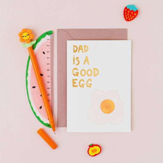 Dad Is A Good Egg Father's Day card featuring a smiling fried egg with gold foil lettering on a contemporary white background.