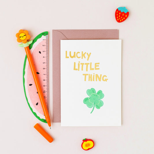 Lucky Little Thing greeting card featuring a smiling four leaf clover with gold foil lettering on a contemporary white background.
