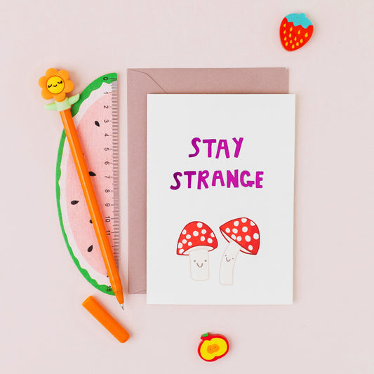 Hand drawn fly agaric mushroom greeting card reading "Stay Strange" with gold foil lettering on a white background.
